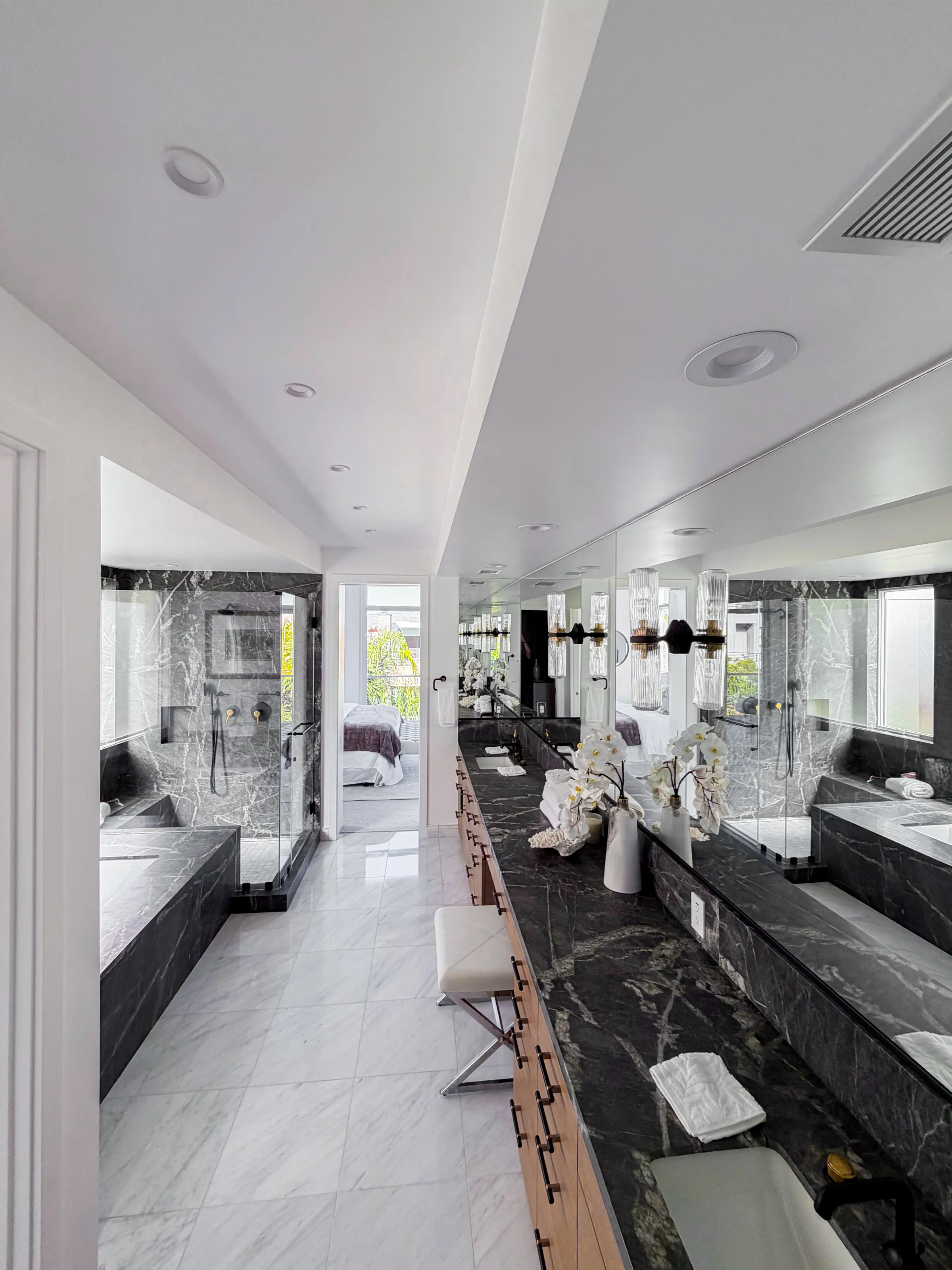 Long primary bathroom with a dark marble vanity, oak cabinetry, a marble-clad tub, a frameless glass shower and white marble floors