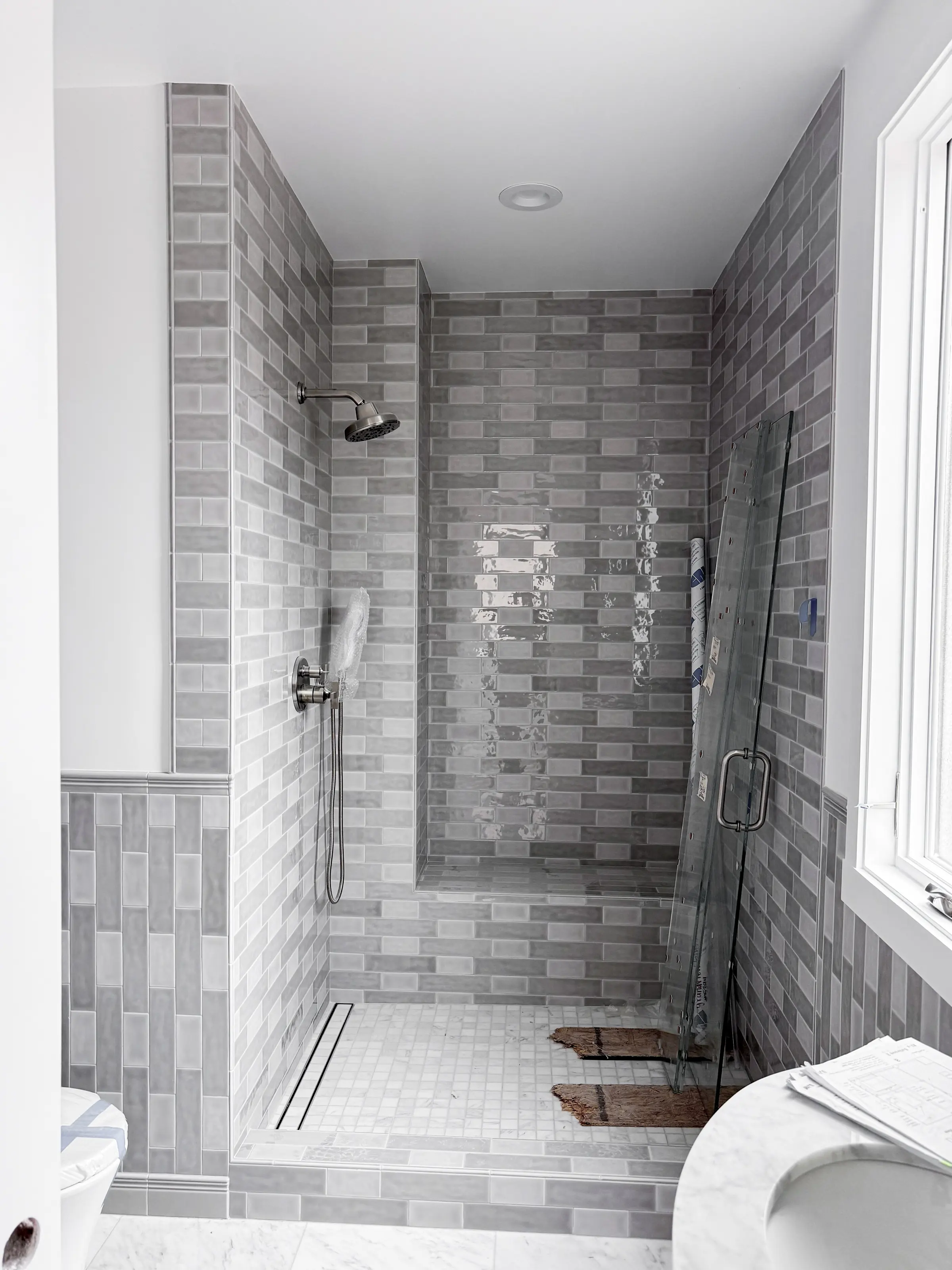 Walk-in shower in glazed grey subway tile with a linear drain and marble mosaic floor, with the glass panel ready to install