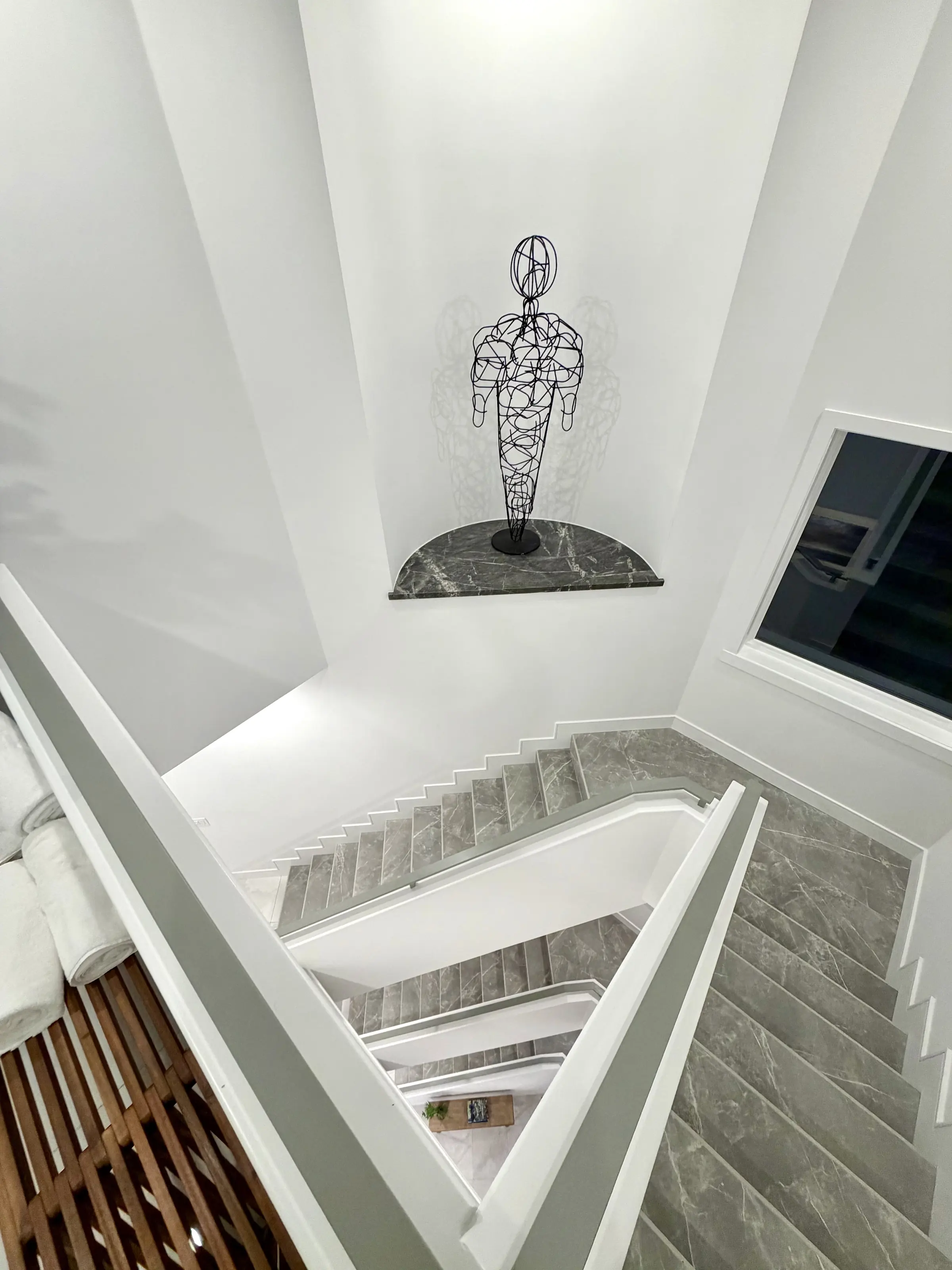 Looking down a white stairwell past a wire figure sculpture set on a marble niche shelf
